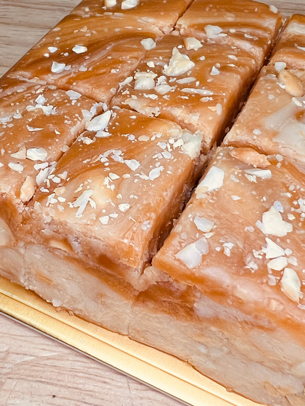 Handmade salted caramel cashew fudge cut into thick squares with a glossy caramel topping and sprinkled with chopped cashews, displayed on a gold tray over a light wooden surface.