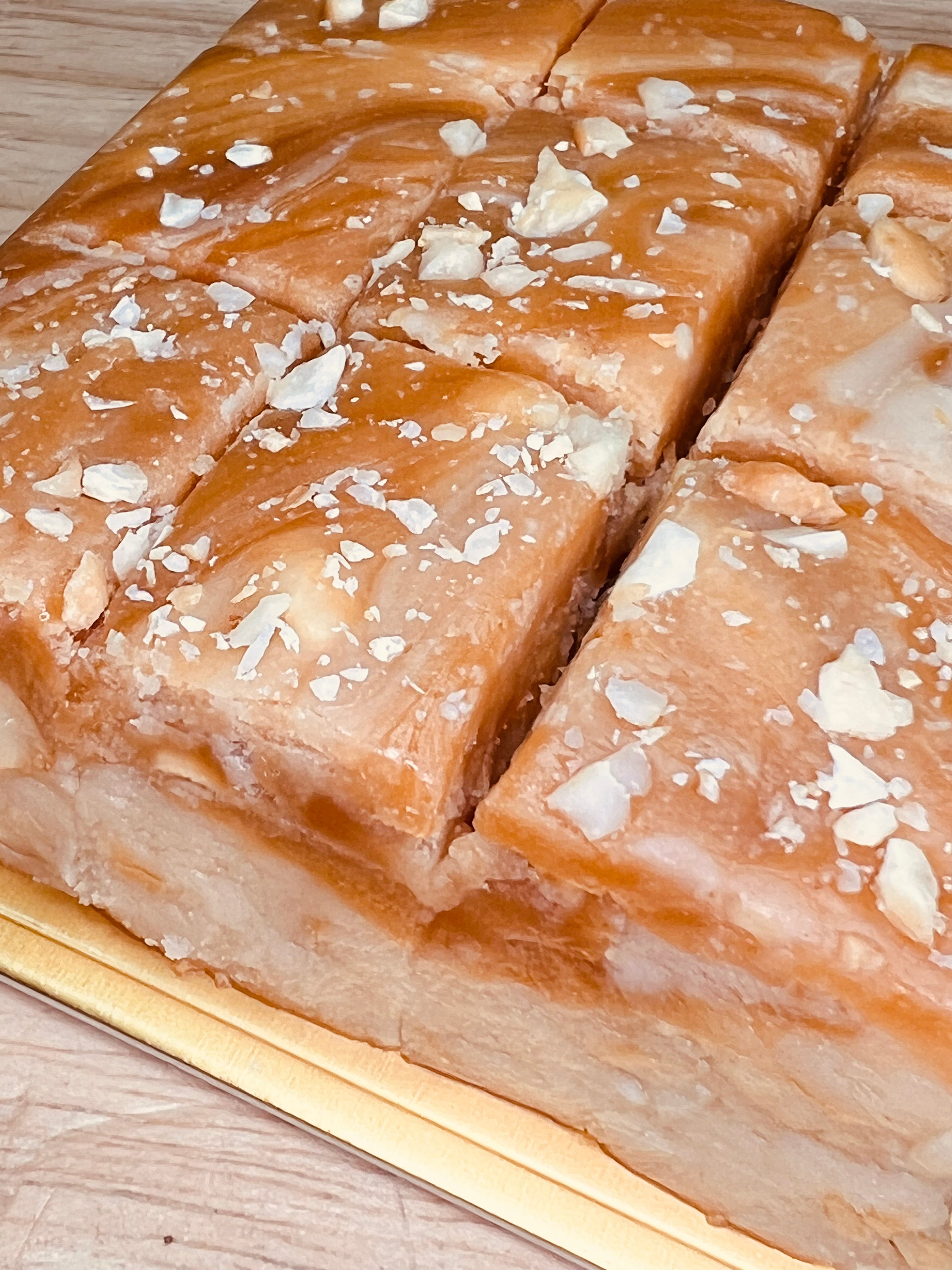 Handmade salted caramel cashew fudge cut into thick squares with a glossy caramel topping and sprinkled with chopped cashews, displayed on a gold tray over a light wooden surface.