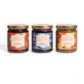 Three jars of In the Curious Kitchen gourmet preserves featuring Strawberry Jalapeño Jam, Bourbon Blueberry Jam, and Bourbon Apple Butter, beautifully arranged on a white background with gold lids and colorful artisan labels.