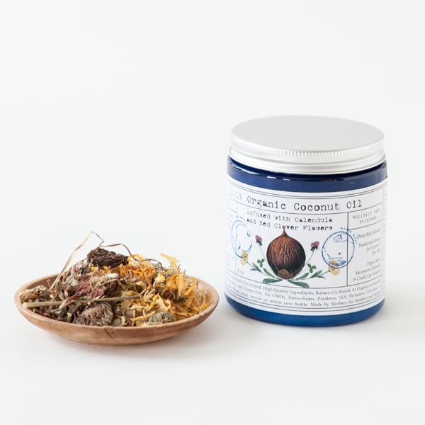 Jar of coconut oil with a bowl of dried herbs on a white background. Infused Organic Coconut Oil with Calendula & Red Clover – 100% Organic Moisturizing Body Oil