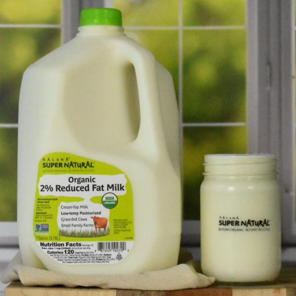 Kalona SuperNatural Organic 2% reduced fat milk in a 1-gallon jug beside a glass jar of cream-top milk on a windowsill, made from grass-fed cows and USDA Organic certified.
