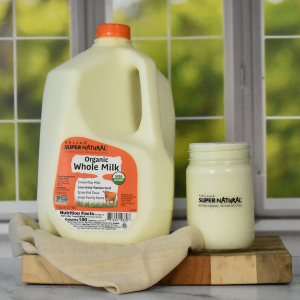 Kalona SuperNatural Organic Whole Milk gallon jug with a glass jar of milk on a wooden board, featuring USDA Organic, grass-fed, low-temperature pasteurized milk from regenerative family farms.