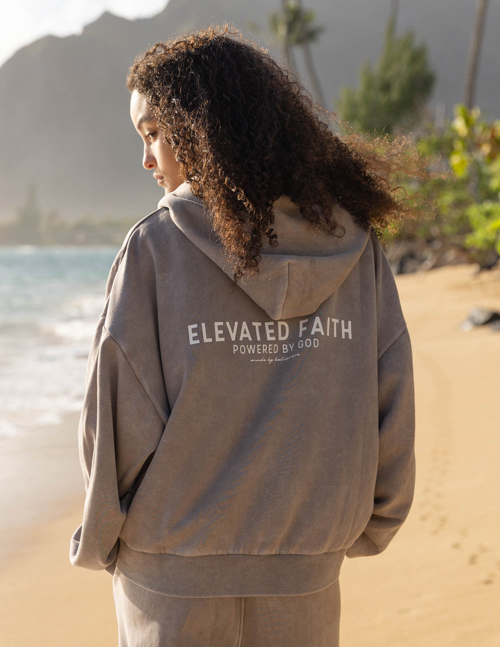Person wearing a hoodie with 'Elevated Faith' text on a beach