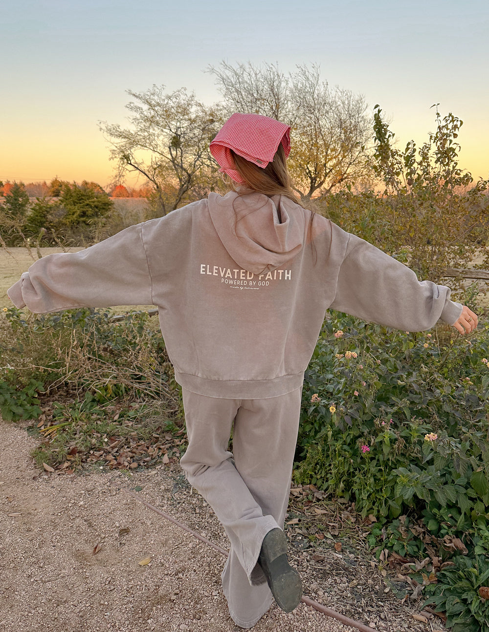 Person wearing a beige 'Elevated Faith' hoodie and pants in a natural setting with a pink hat.