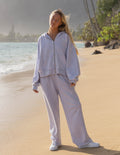 A woman stands on a sandy beach wearing a matching light lavender zip-up hoodie and wide-leg sweatpants set, paired with white sneakers. The background shows the ocean, shoreline, and palm trees in soft, warm light.