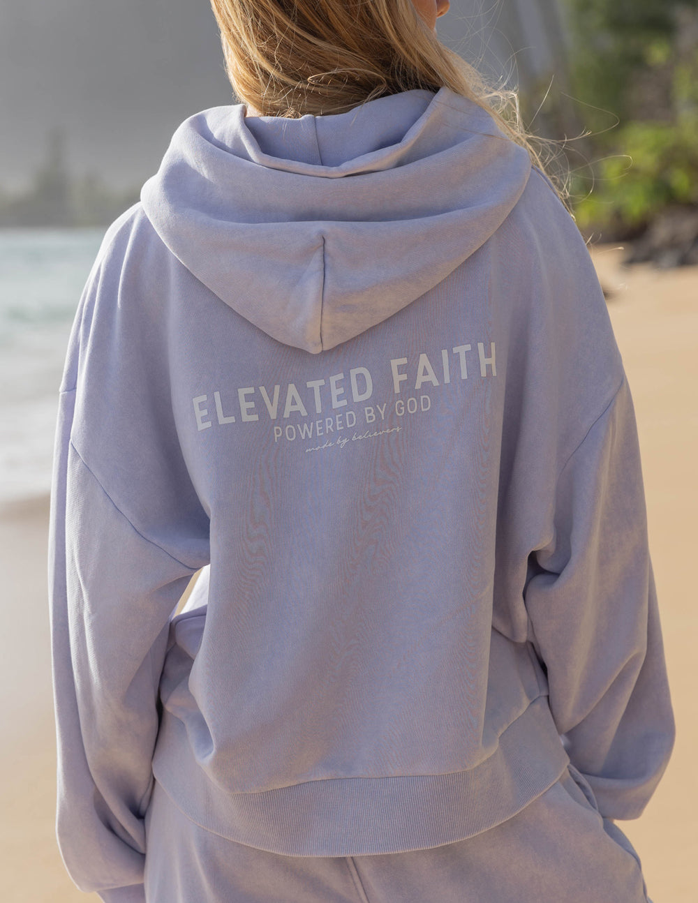 Person wearing a light blue hoodie with 'Elevated Faith' text on the beach