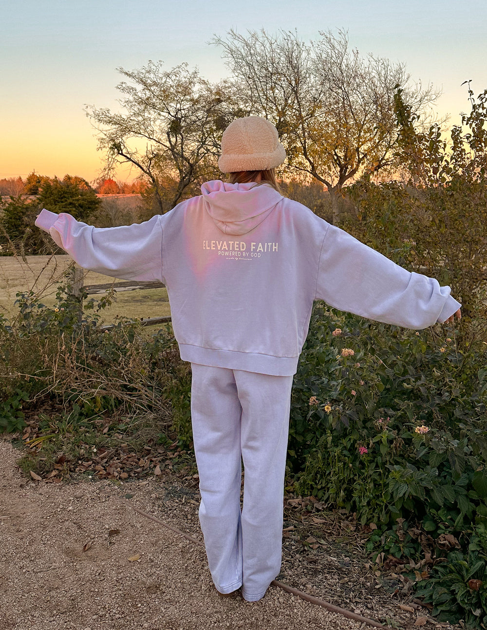 Person wearing a white hoodie and pants with 'Elevated Faith' text, standing outdoors with arms outstretched.