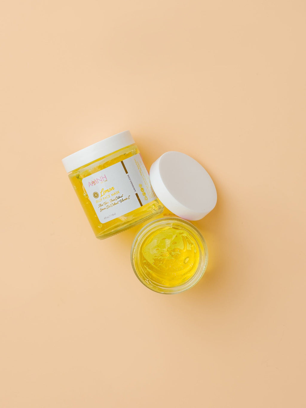 Two jars of yellow cream with white lids on a beige background. AMINNAH Lemon Jelly Face Mask with open and closed jars displaying the bright yellow jelly formula. Vegan, cruelty-free clean beauty face mask made with natural ingredients including Aloe Vera, Rose Extract, Vitamin E, Coconut Oil, and Lemon Zest Extract from a female-owned brand.