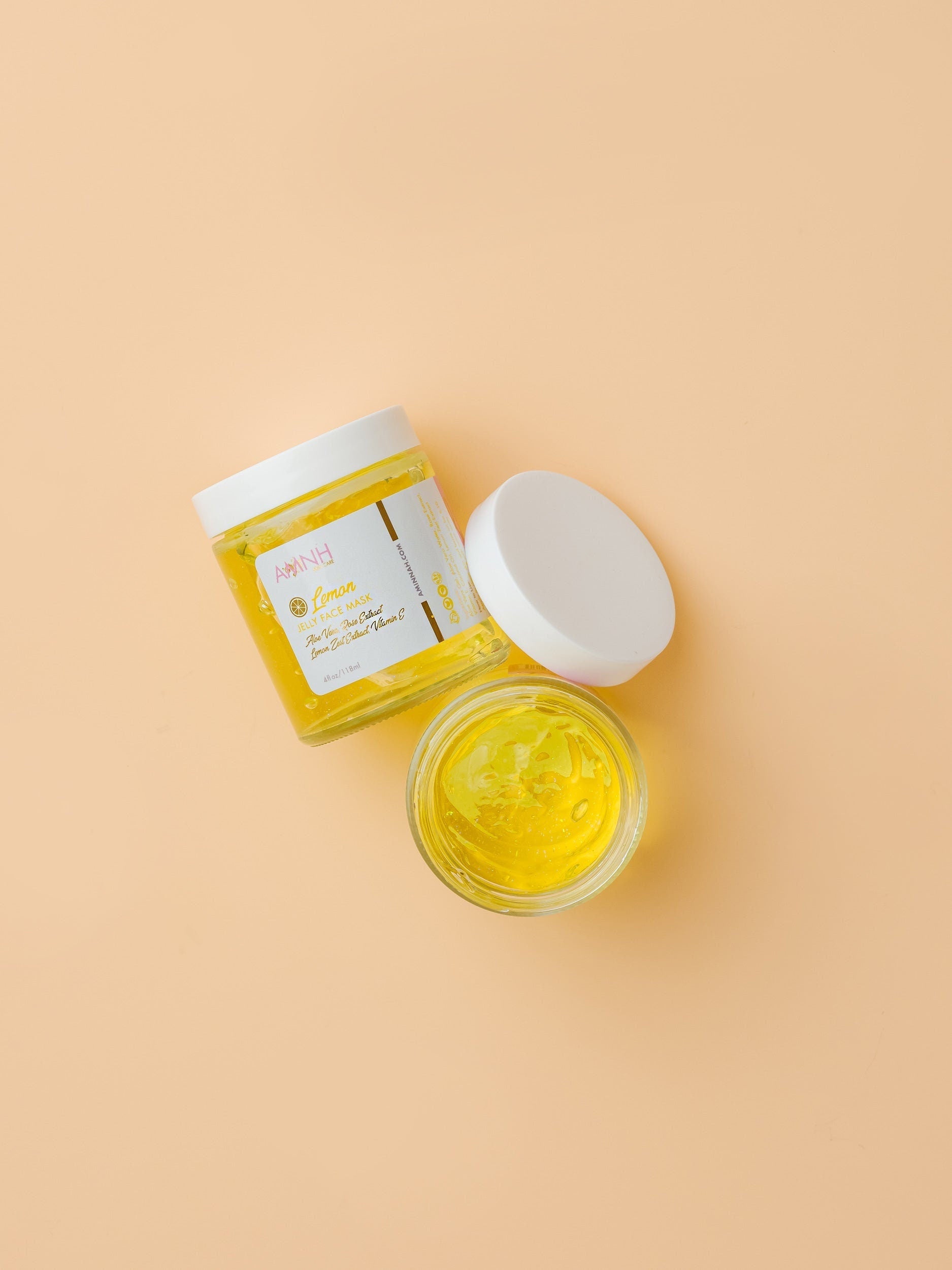 Two jars of yellow cream with white lids on a beige background. AMINNAH Lemon Jelly Face Mask with open and closed jars displaying the bright yellow jelly formula. Vegan, cruelty-free clean beauty face mask made with natural ingredients including Aloe Vera, Rose Extract, Vitamin E, Coconut Oil, and Lemon Zest Extract from a female-owned brand.