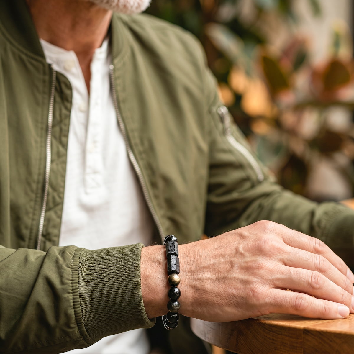 A close-up of a man's hand resting on a wooden table, wearing a black beaded bracelet with polished stone accents. He is wearing an olive green bomber jacket over a white henley shirt, with a softly blurred indoor background. Direction of Wealth & Success - The Spiritual Protection Bracelet with Black Tourmaline and Golden Obsidian for Absorbing Negativity