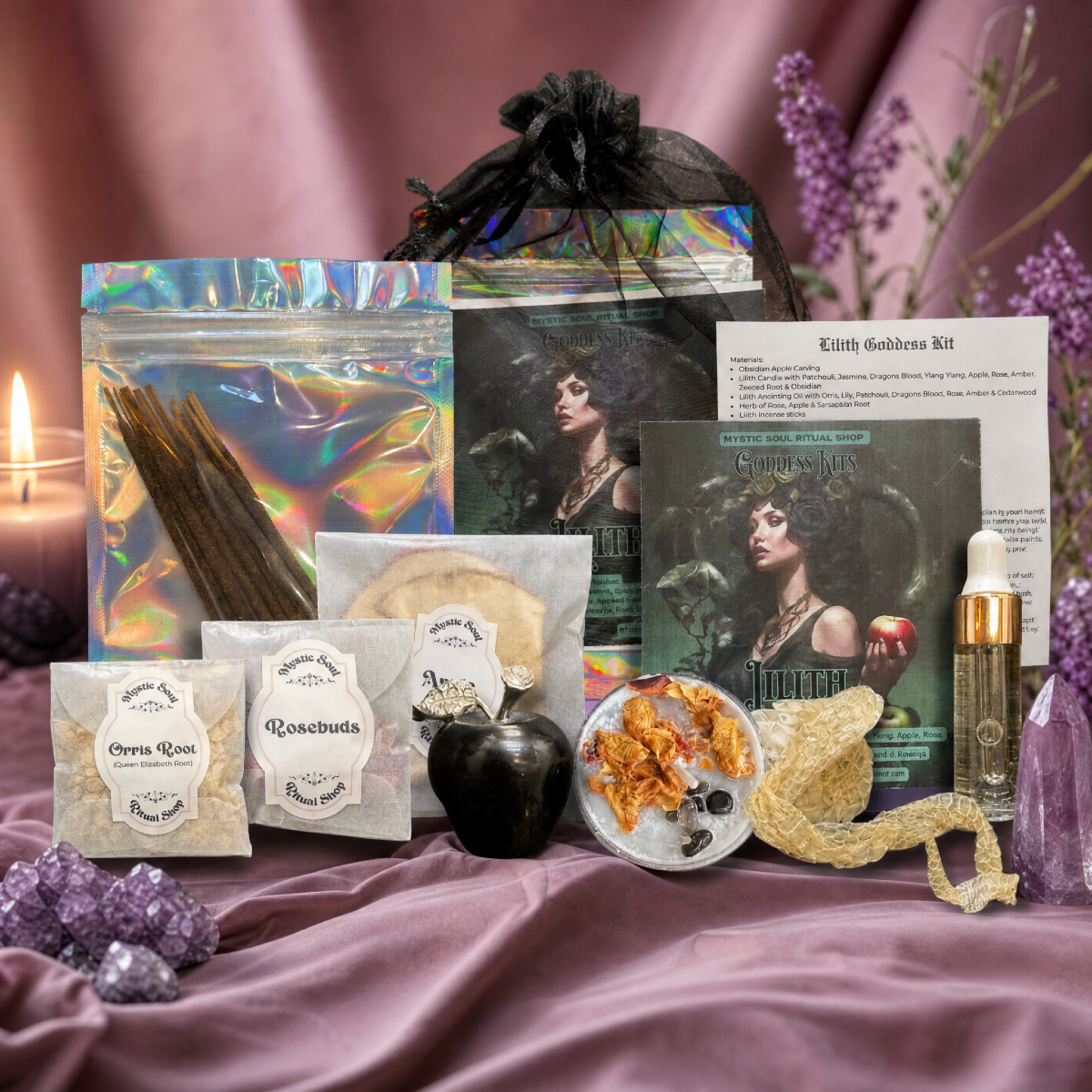 A styled product display of a 'Kith Goddess Kit' containing various ingredients and accessories, including raw rose petals, cinnamon sticks, dried herbs, a black apple, crystals, and a small glass bottle, arranged on a draped mauve satin fabric with purple flowers and a lit candle. Metaphysical items. Lilith Devotional Goddess Kit with orris root, rosebuds, incense sticks and oil for evoking Lilith: Goddess of independence, sexuality, feminine sovereignty, and righteous rage. Dark feminine occult work.
