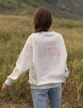 A person standing in a grassy outdoor field, viewed from behind, wearing a cream sweatshirt with a pink graphic that reads 'LOST AND FOUND' and light blue jeans.