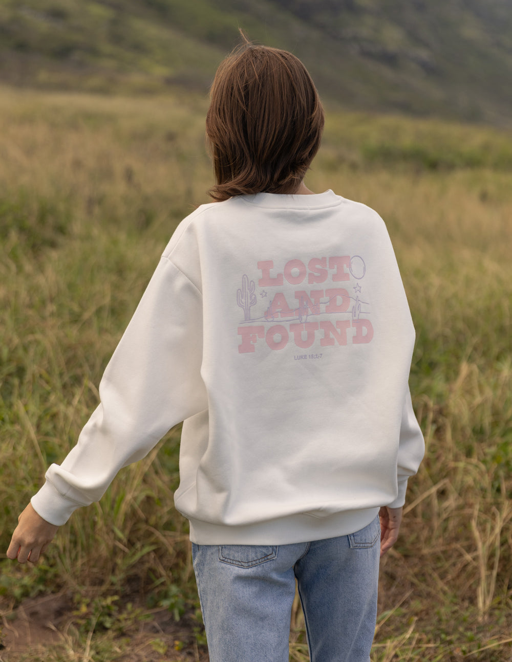A person standing in a grassy outdoor field, viewed from behind, wearing a cream sweatshirt with a pink graphic that reads 'LOST AND FOUND' and light blue jeans.