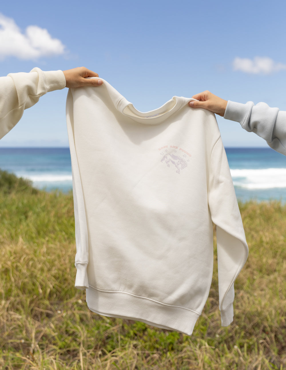 Two people holding a white sweatshirt against a scenic background of grass and ocean.