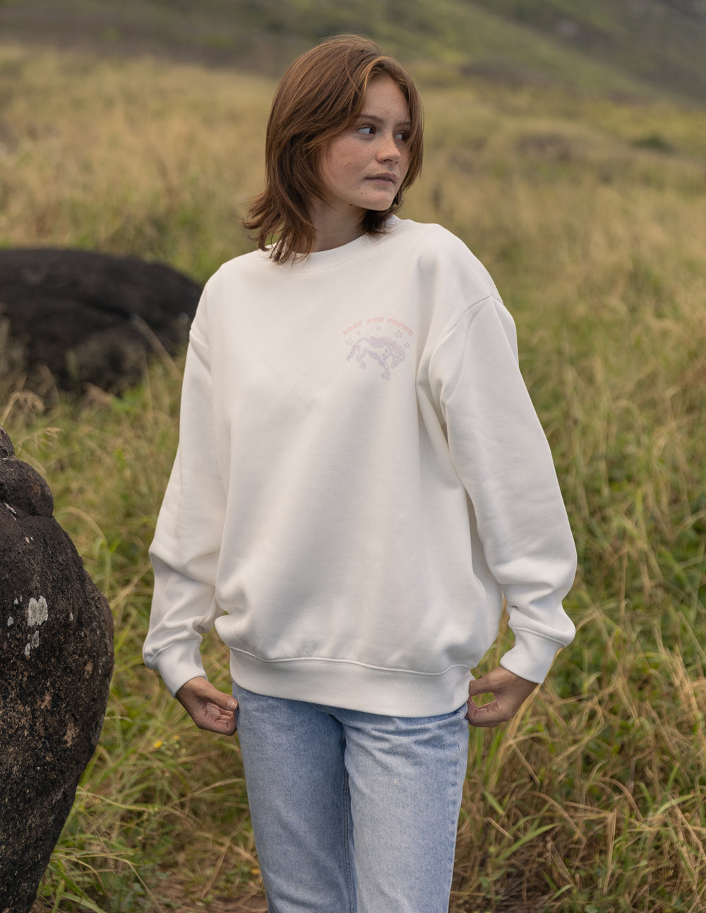Person wearing a white sweatshirt and light blue jeans standing in a grassy field with animals in the background.