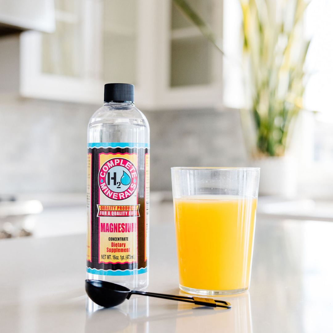 Bottle of Complete Minerals magnesium supplement next to a glass of orange juice on a kitchen counter. Bottle of Complete Essentials Magnesium supplement with benefits listed on a light blue background. Magnesium Supports: strong bones, joints and teeth, heart benefits, dreamy nights, energized muscles, increased energy, stress relief, complete H20 minerals, quality products for a quality life, magnesium concentrate dietary supplement, NET WT. 16oz. 1pt 473 mL