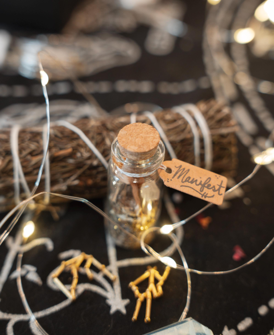 Small glass bottle with cork lid on a dark surface with string lights and decorative elements. Manifest Spell Jar