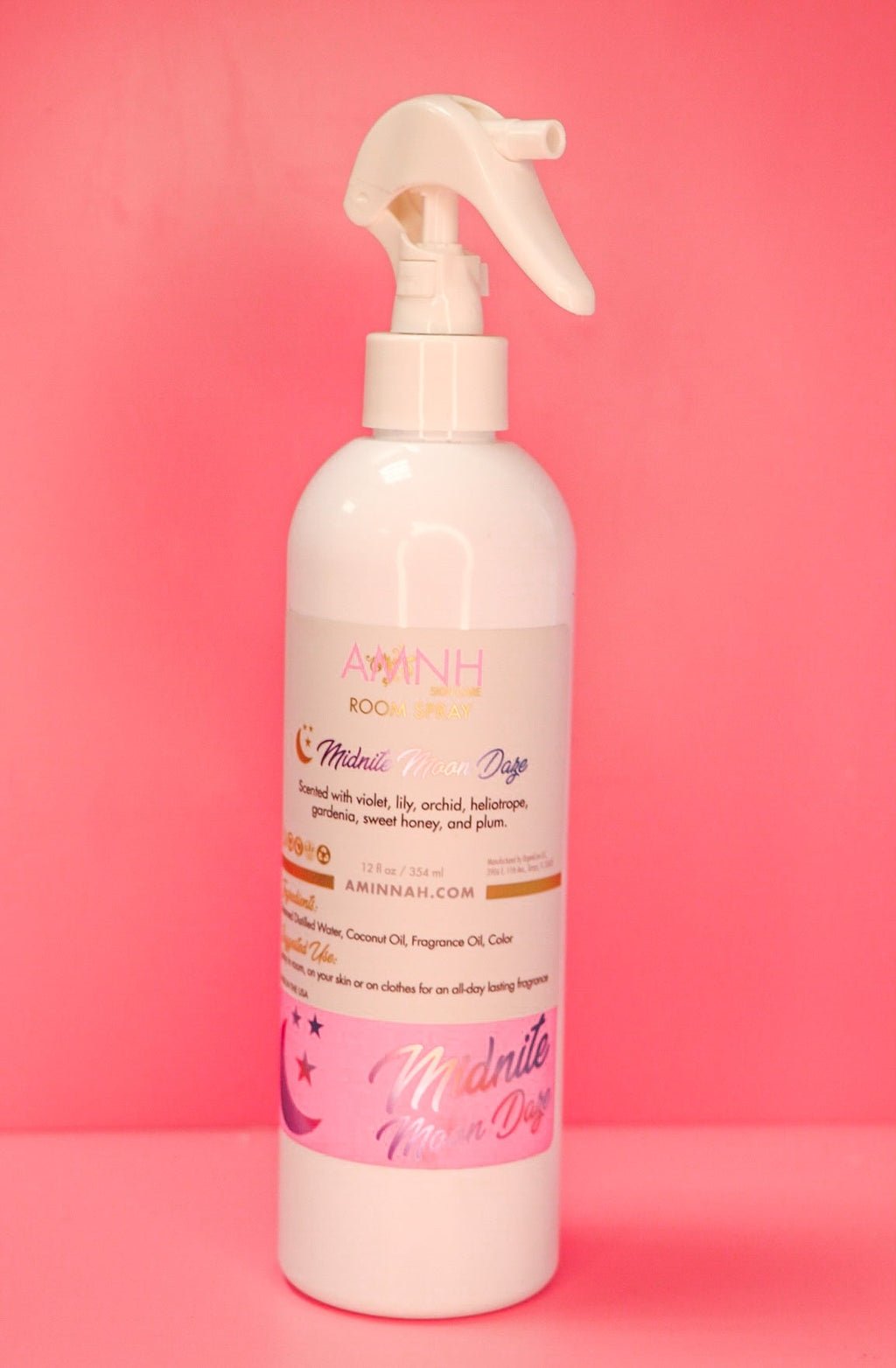 AMINNAH Midnite Moon Daze Room Spray in a white spray bottle displayed on a pink background. Clean beauty, cruelty-free, vegan home fragrance spray crafted with naturally derived ingredients including steamed distilled water, coconut oil, and fragrance oil. Features enchanting notes of violet, lily, orchid, heliotrope, gardenia, sweet honey, and plum to refresh rooms, linens, and fabrics. Female-owned brand available at Tree of Life Grocers.