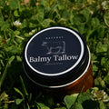 A jar of Balmy Tallow natural hand and foot cream resting on green grass with clover. The jar has a black lid with a white circular border and a simple line drawing of a cow, with the brand name 'Balmy Tallow' printed in white.