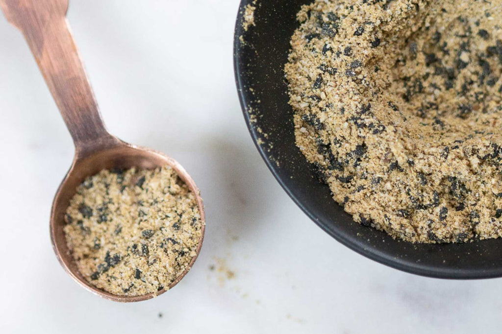 Close-up overhead view of a black bowl filled with Caesar salad seasoning blend featuring herbs, spices, Parmesan-style seasoning, and black pepper, with a wooden spoon holding a scoop on a light marble surface.