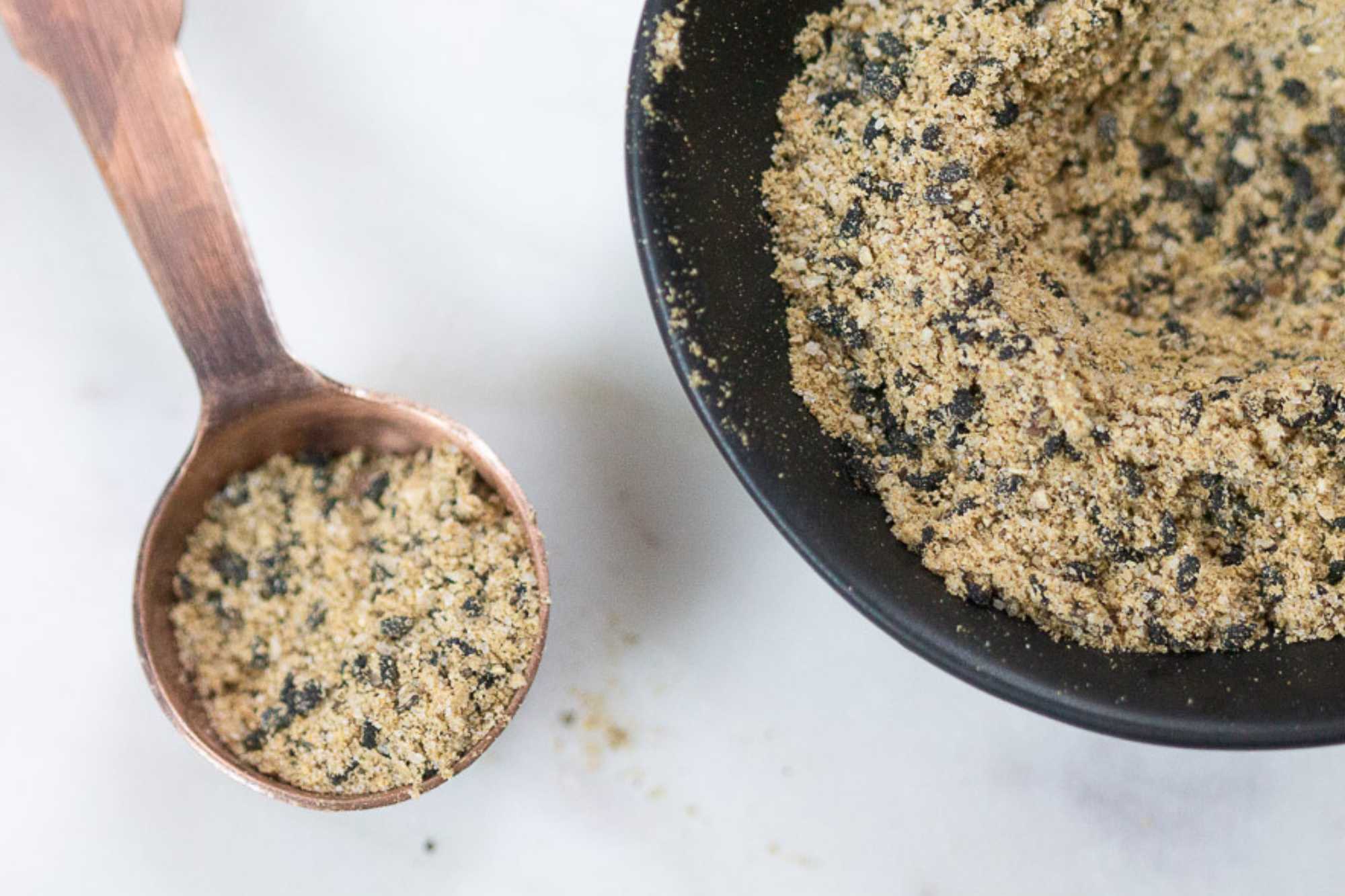 Close-up overhead view of a black bowl filled with Caesar salad seasoning blend featuring herbs, spices, Parmesan-style seasoning, and black pepper, with a wooden spoon holding a scoop on a light marble surface.