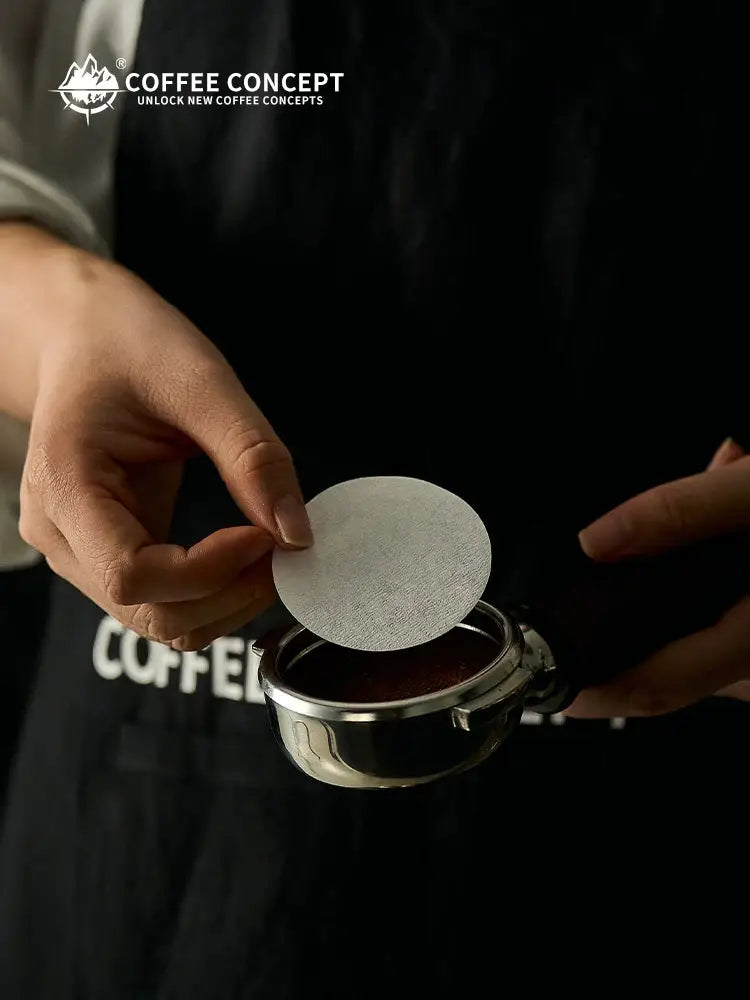 Person placing a round espresso puck screen filter paper over a portafilter basket filled with ground coffee, Coffee Concept branded barista accessory.