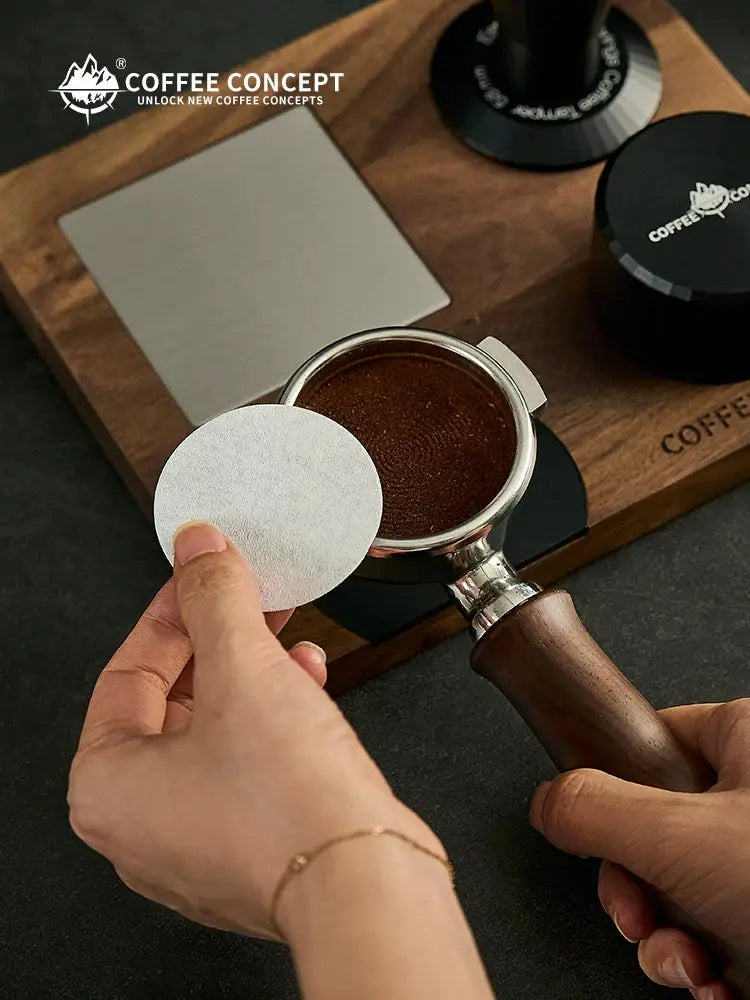Espresso puck screen filter paper being placed over freshly ground coffee in a wooden-handle portafilter, designed for 51mm, 53/54mm and 58mm espresso baskets to help reduce coffee grounds buildup, improve water distribution and make basket cleanup easier; disposable round paper espresso filter accessory for home baristas, coffee lovers and espresso machine brewing.