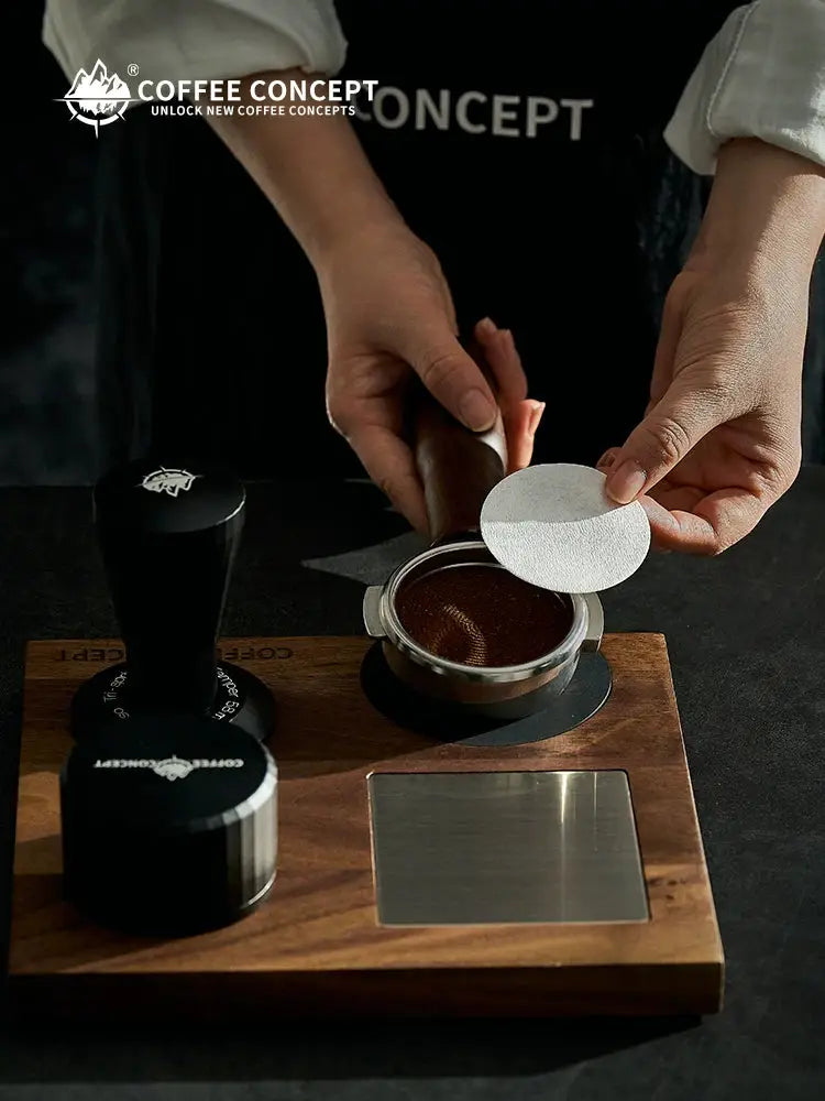 Barista preparing espresso with round puck screen filter paper over freshly ground coffee in a portafilter basket, Coffee Concept espresso accessory for 51mm, 53/54mm and 58mm baskets, disposable coffee filter paper for cleaner puck removal, reduced coffee grounds buildup, easy basket cleanup and home espresso brewing.