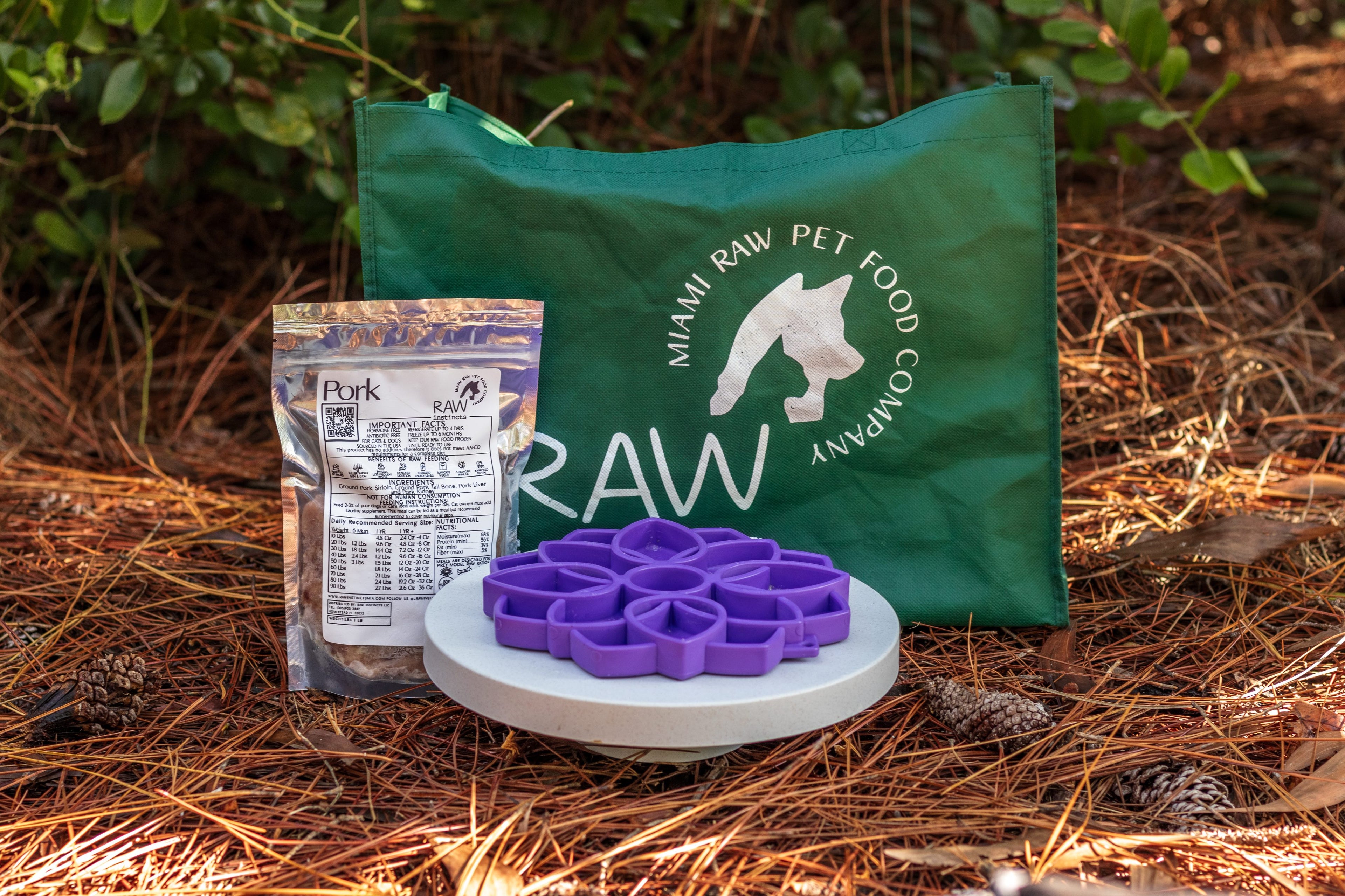 Raw PMR pork meal blend package with a purple flower-shaped slow feeder and Miami Raw Pet Food Company tote bag displayed outdoors on a natural forest floor, showcasing a biologically appropriate raw diet for cats and dogs.