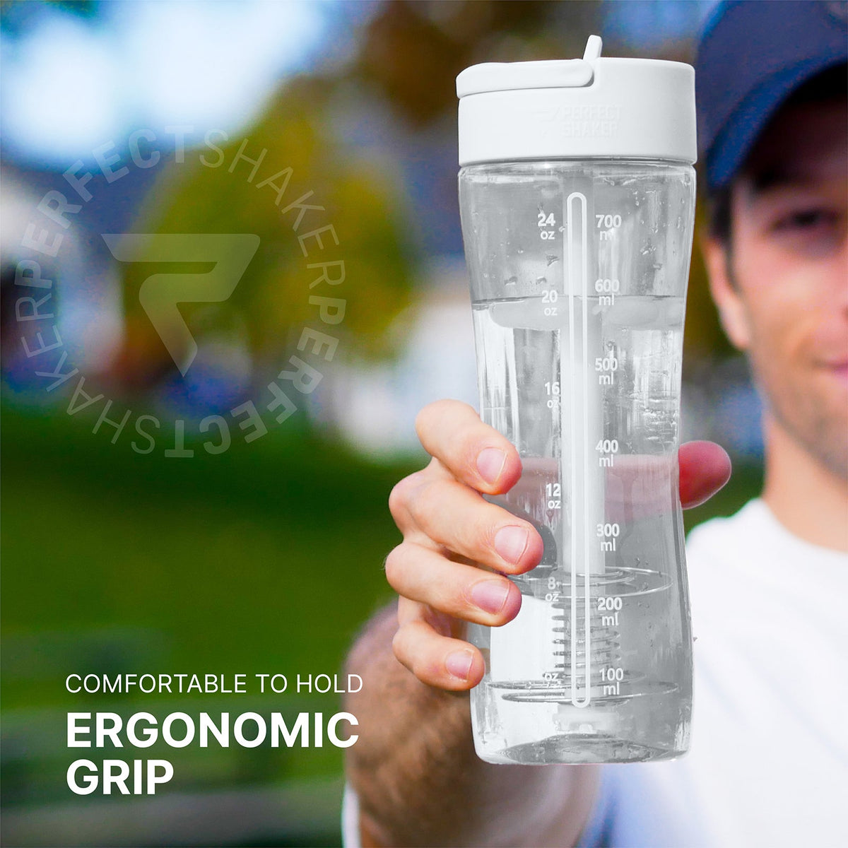 A man holding a clear plastic water bottle with a white lid and measurement markings. The image features a blurred outdoor background and text overlays that read 'COMFORTABLE TO HOLD ERGONOMIC GRIP' and a circular watermark 'SHAKERSPERFECT'.