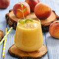 A glass of smooth, creamy yellow beverage, likely a mango or peach smoothie, garnished with a small mint sprig, set on a rustic wood slice coaster. Fresh peaches and additional wooden coasters are arranged on a light blue wooden table, with two yellow-and-white striped straws on the left. Peaches & Cream Smoothie Mix