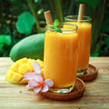 Two glasses of creamy pineapple mango smoothie with reusable bamboo straws, fresh pineapple and mango slices, mint garnish, and tropical flowers on a rustic wooden table, set against a lush green island-inspired background.