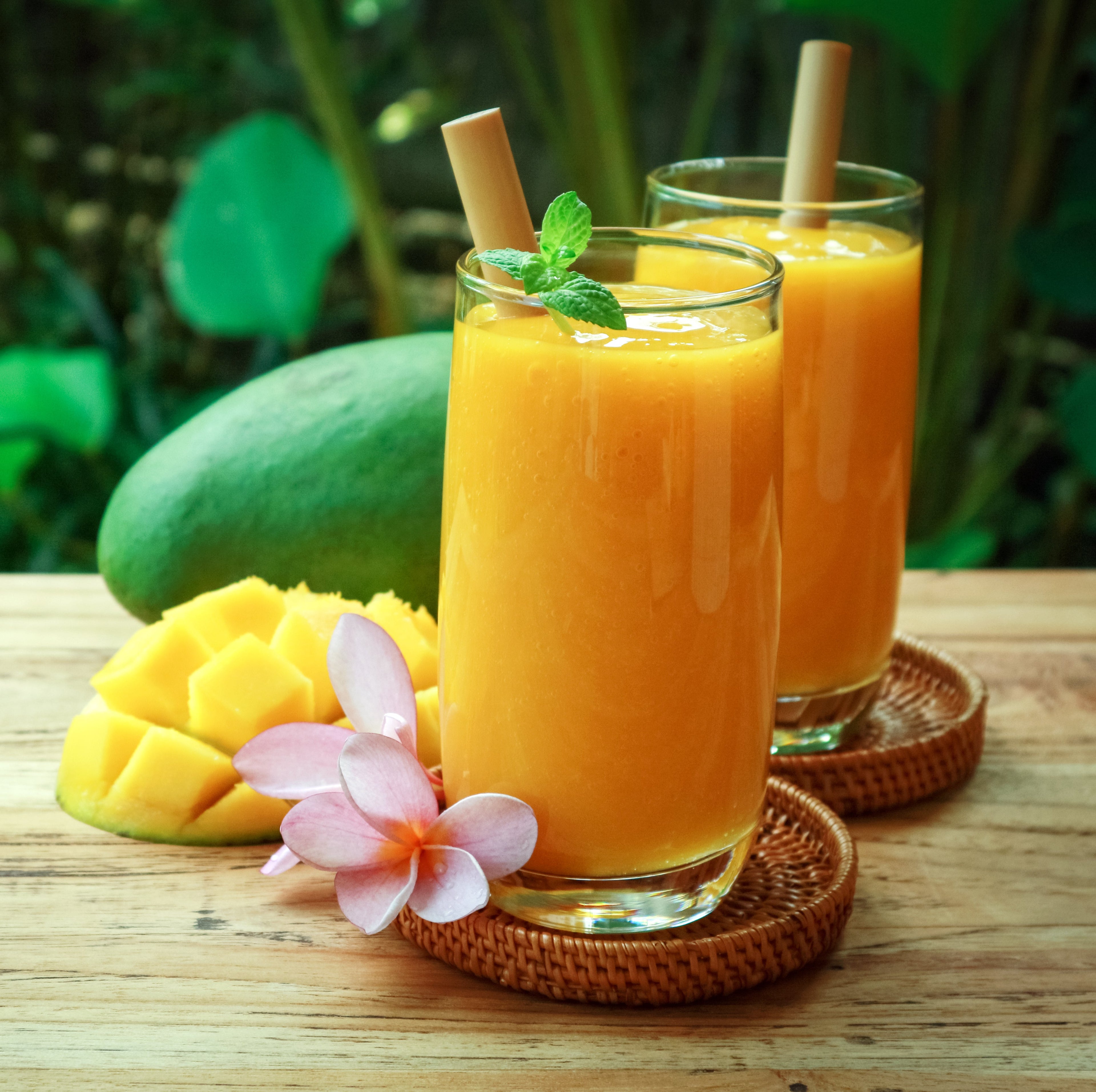 Two glasses of creamy pineapple mango smoothie with reusable bamboo straws, fresh pineapple and mango slices, mint garnish, and tropical flowers on a rustic wooden table, set against a lush green island-inspired background.