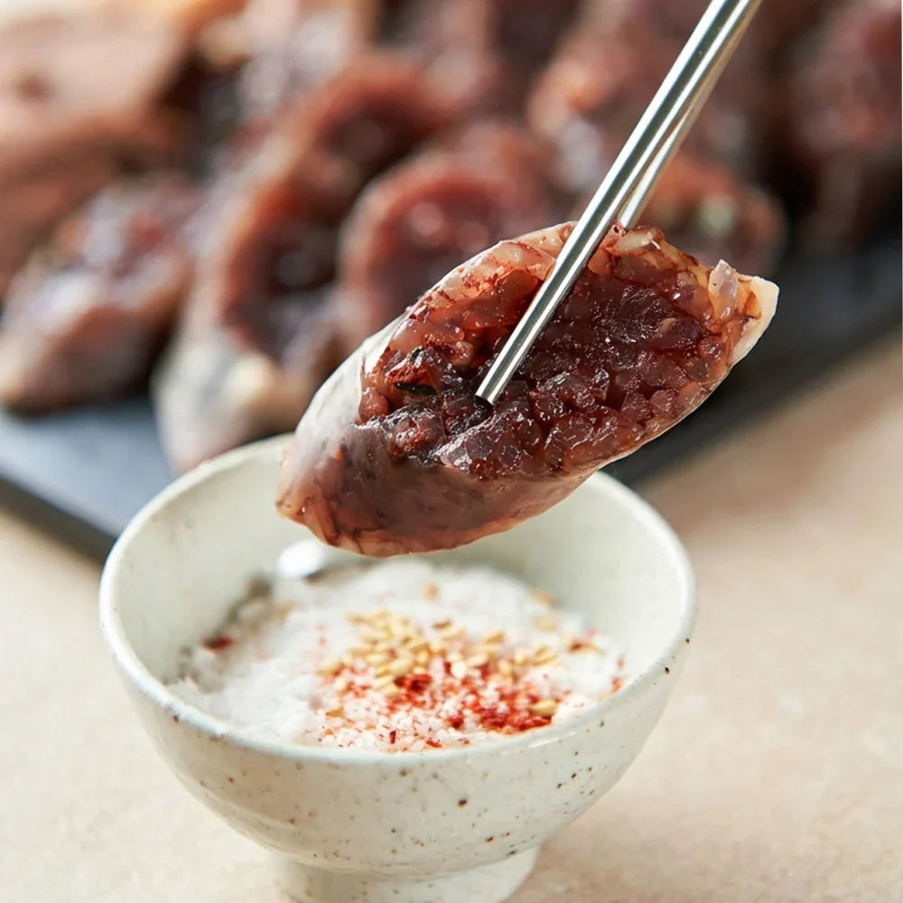 Premium Korean blood sausage (sundae) filled with glutinous rice, held with stainless steel chopsticks and dipped into a bowl of seasoned salt with sesame seeds and red pepper flakes.