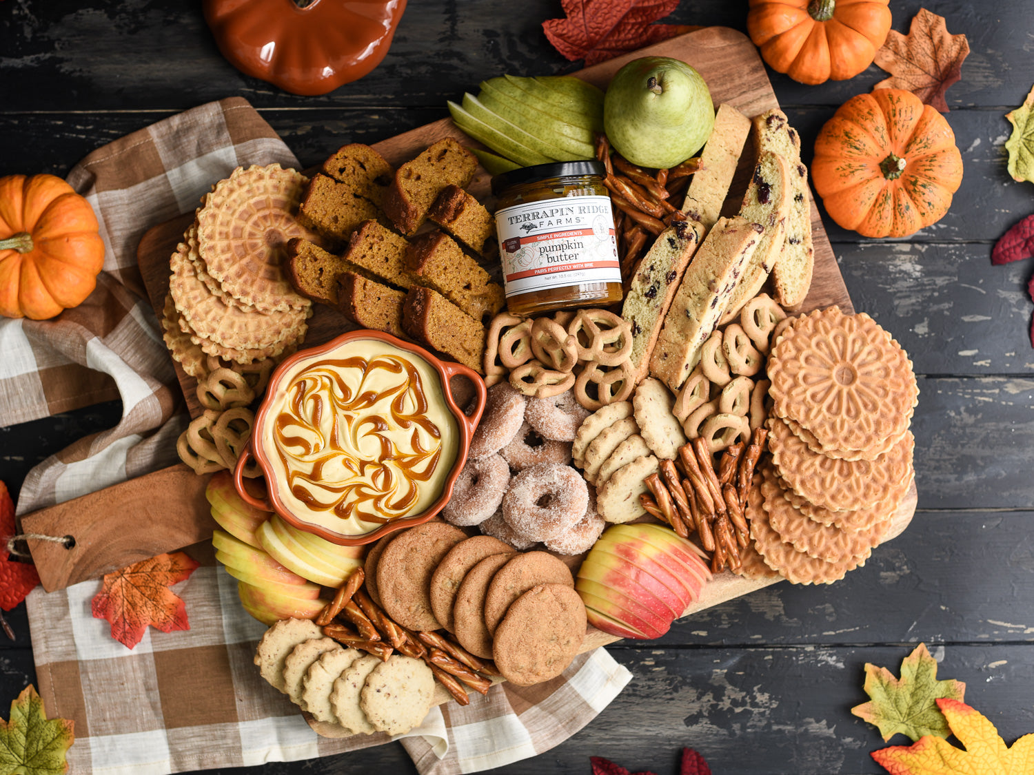 Autumn charcuterie board featuring pumpkin butter, sliced apples, cookies, pretzels, biscotti, and seasonal treats arranged on a rustic wooden serving board with pumpkins and colorful fall leaves.