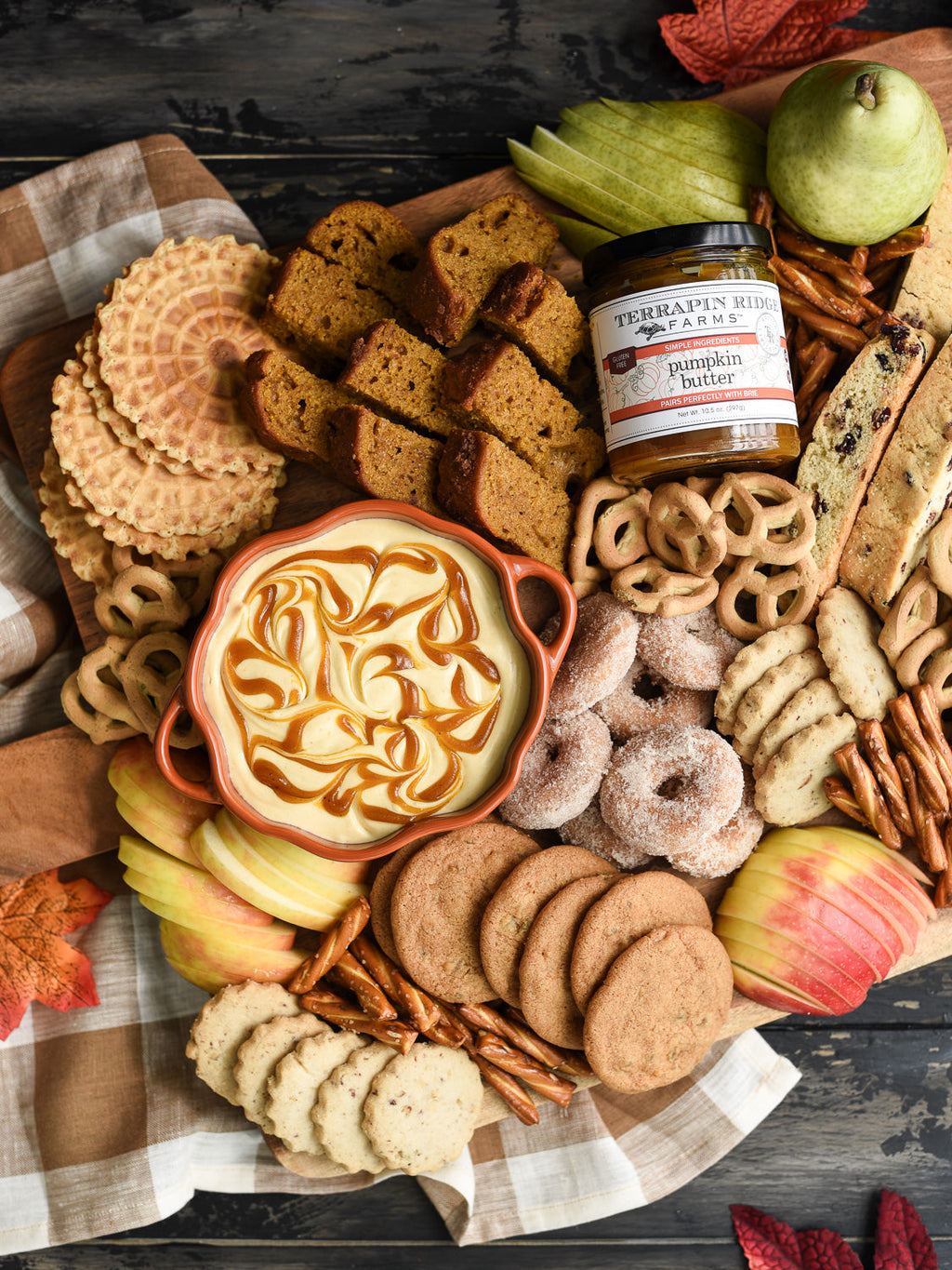 Autumn charcuterie board featuring creamy pumpkin dip, Terrapin Ridge Farms Pumpkin Butter, sliced apples, pumpkin bread, cookies, pretzels, donuts, and biscotti arranged on a rustic wooden serving board for a cozy fall gathering.