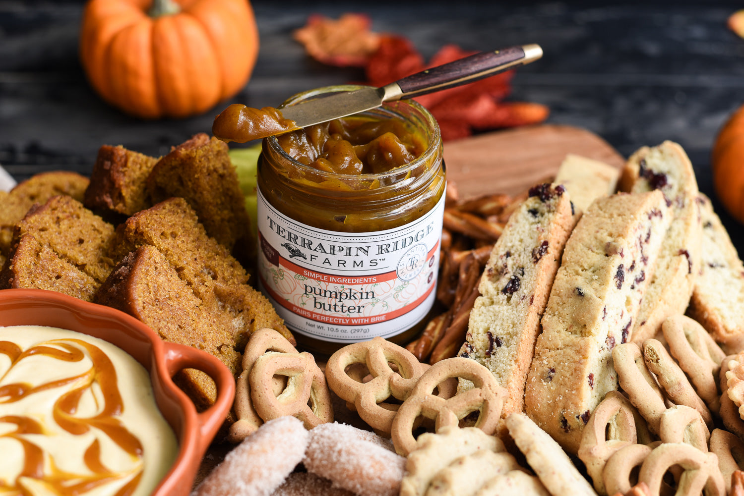 Terrapin Ridge Farms Pumpkin Butter served on a festive fall charcuterie board with pumpkin bread, biscotti, pretzels, cookies, caramel dip, pumpkins, and autumn leaves, showcasing a cozy seasonal entertaining spread.