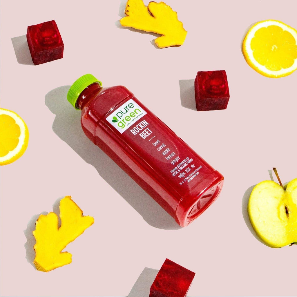 Red juice bottle labeled 'pure green' surrounded by fruit pieces on a light pink background. Cold-Pressed juice drink with Beets, Carrot, Apple, Lemon and Ginger. A NON-GMO juice drink in 16 - oz bottle. The Pure Green Cold Pressed Juice company is a proud supporter of local and organic farms.