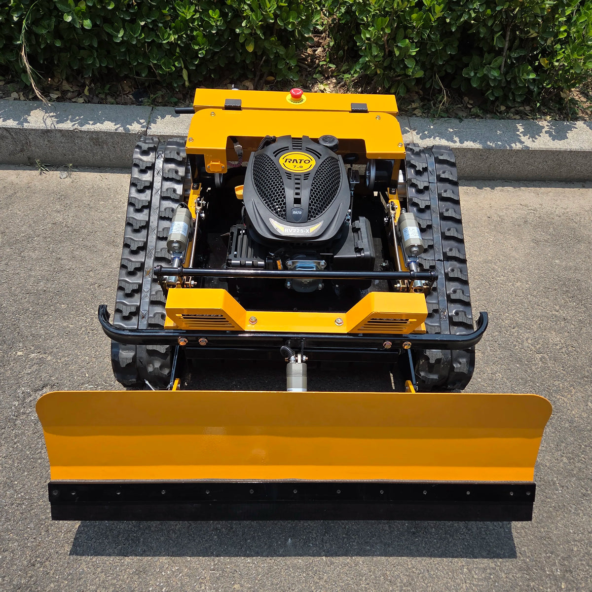 A yellow and black tracked compact bulldozer with a large front blade, photographed from above on an asphalt surface with green bushes in the background. The engine cover shows the brand 'RATO'.