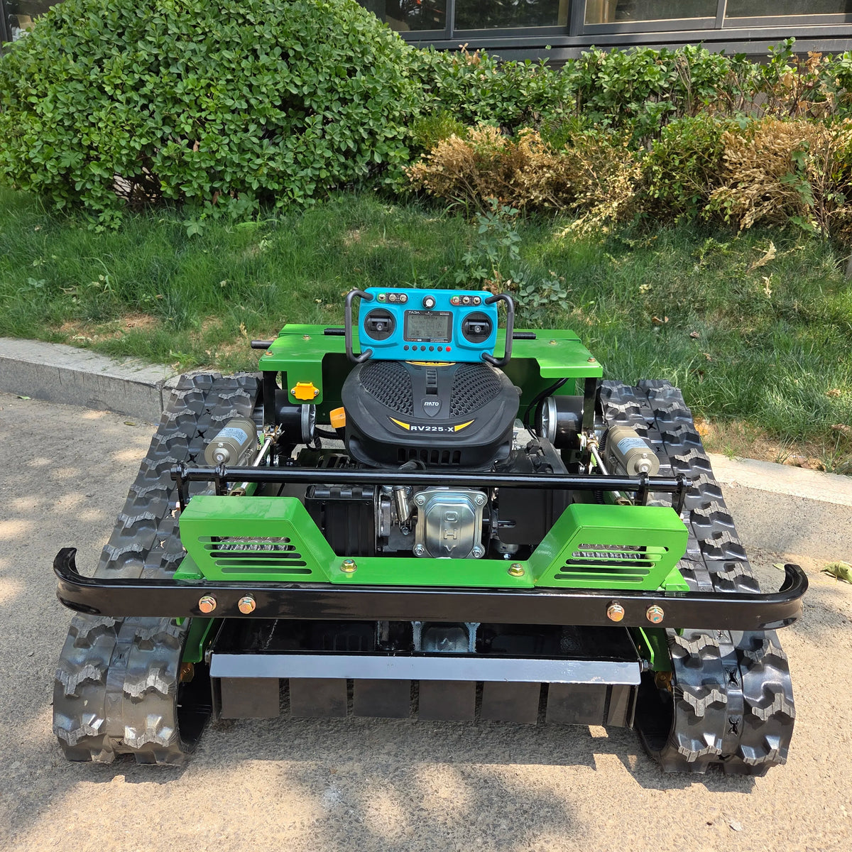 A green and black tracked lawn mower with a blue control panel and engine cover, photographed outdoors on a paved surface with grass and bushes in the background.