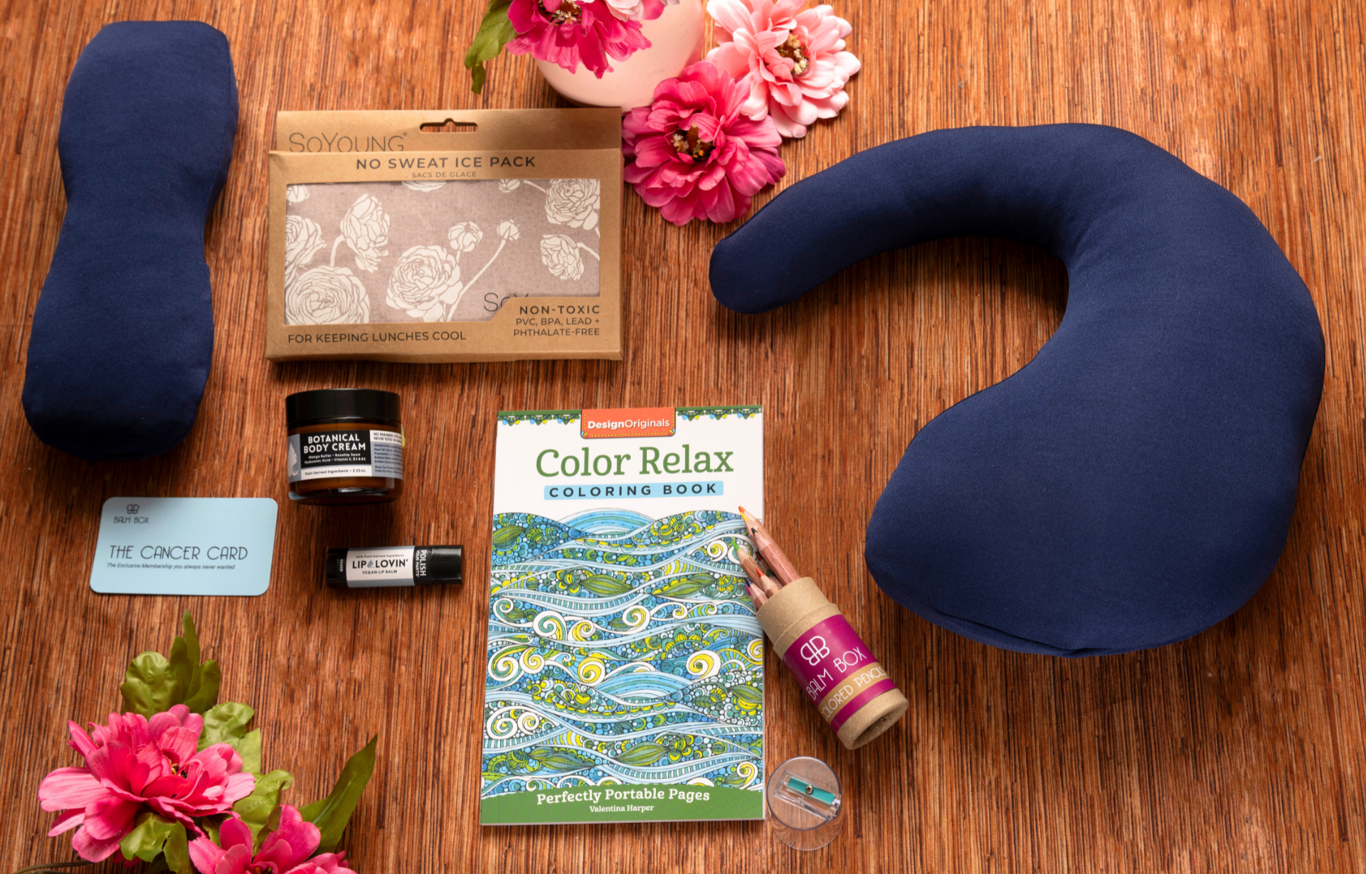 The Best Mastectomy Care Package featuring a blue travel neck pillow, cooling ice pack, relaxing coloring book, lip balm, botanical body cream, and thoughtful self-care essentials arranged with pink flowers on a warm wooden background — breast cancer recovery gift, mastectomy recovery care package, cancer survivor gift, women’s wellness gift box, thoughtful cancer care package, post-surgery comfort gift, self-care gift for cancer patients, recovery gift basket, supportive gift for her, Tree of Life Grocers.