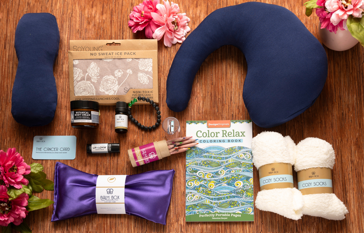 The Best Mastectomy Care Package featuring a navy travel neck pillow, soothing ice pack, cozy socks, satin sleep mask, body cream, lip care, aromatherapy, coloring book, wellness essentials, and thoughtful cancer recovery gifts arranged on a warm wood surface with fresh pink flowers