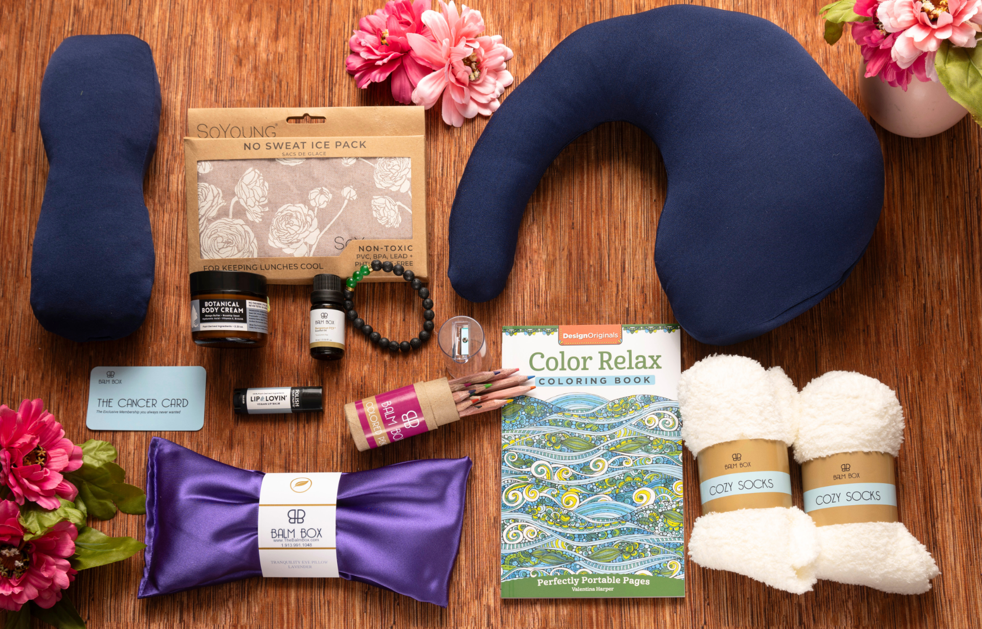 The Best Mastectomy Care Package featuring a thoughtfully curated collection of women’s cancer care, breast cancer recovery, mastectomy recovery, post-surgery comfort, self-care, wellness, relaxation, and survivor support gifts including a soothing neck pillow, sleep eye mask, no-sweat ice pack, cozy socks, body cream, lip care, coloring book, colored pencils, inspirational cancer card, calming aromatherapy oil, and satin sleep accessories on a warm wood background with beautiful pink flowers.
