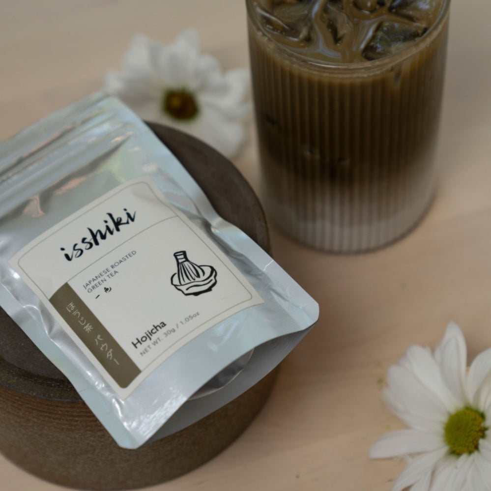 Packaged green tea with 'ishiki' branding next to a glass of iced coffee on a wooden surface.