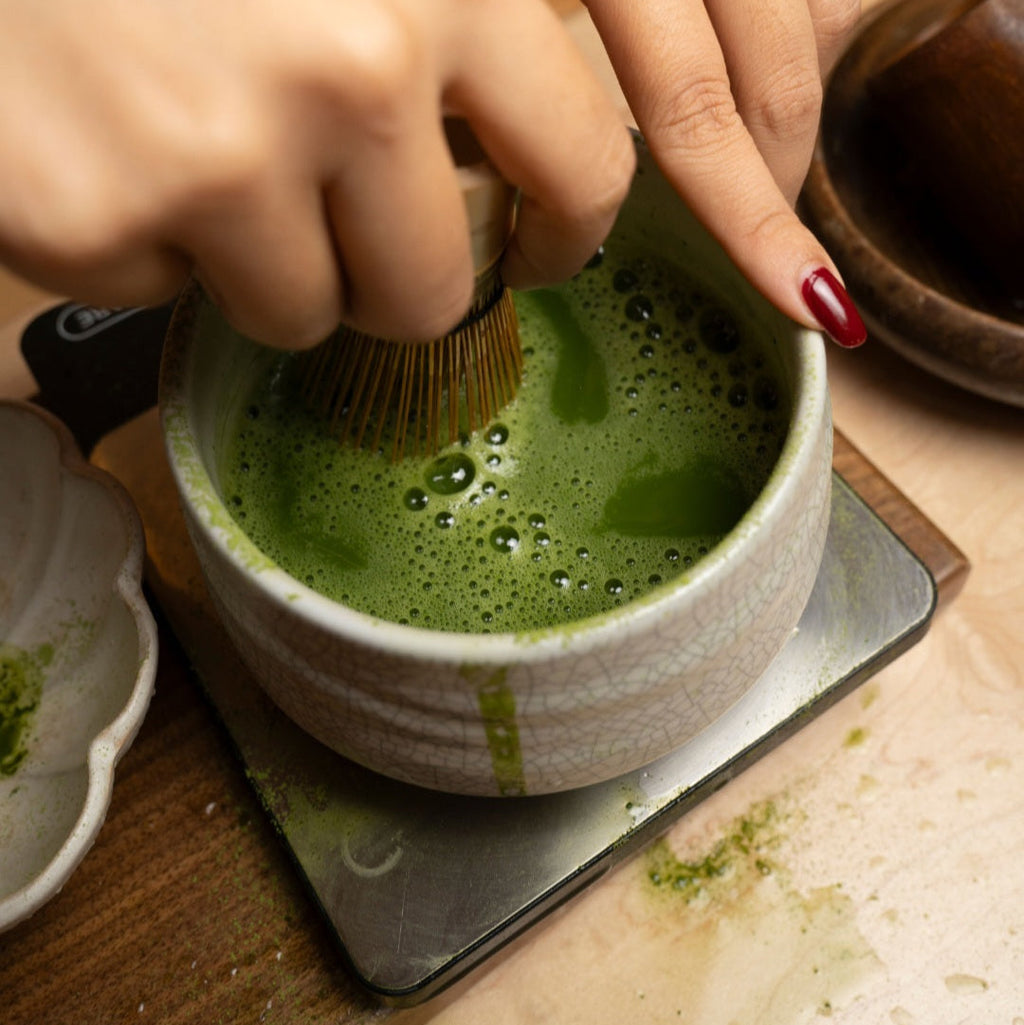 Person using a whisk to prepare matcha green tea in a ceramic bowl.