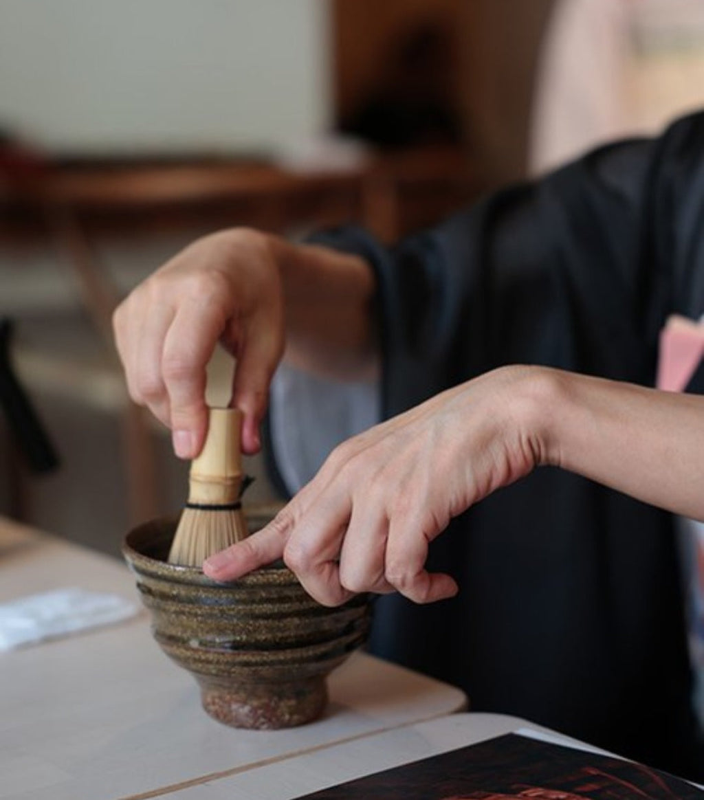 Person using a wooden whisk to prepare matcha in a ceramic bowl.