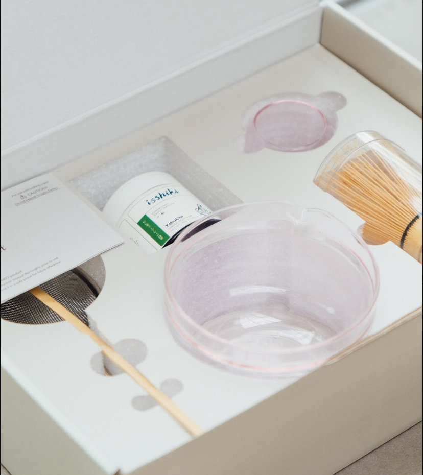 Tea-making set with a pink bowl, whisk, and container in a box.