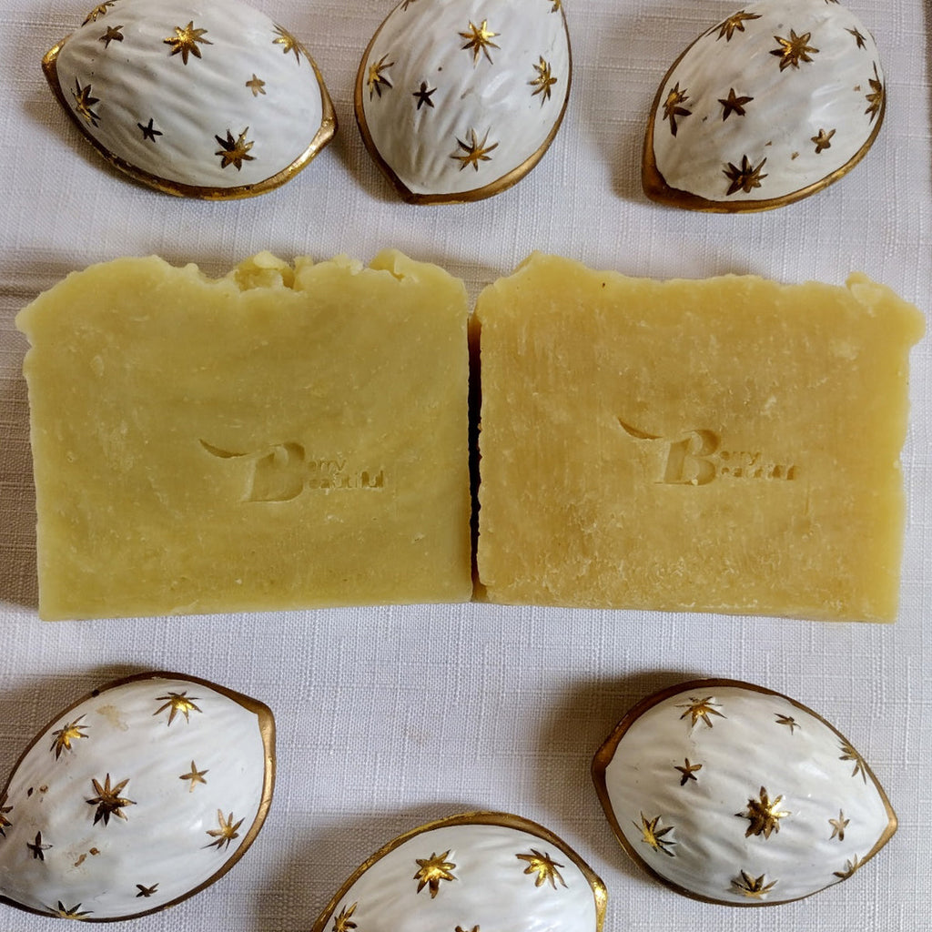 Two bars of soap with a brand logo, surrounded by decorative ceramic containers on a textured surface.