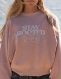 A close-up of a person wearing a dusty pink crewneck sweatshirt with a white graphic that reads 'STAY ROOTED' above three smiling flowers. The background shows a blurred outdoor scene with water.