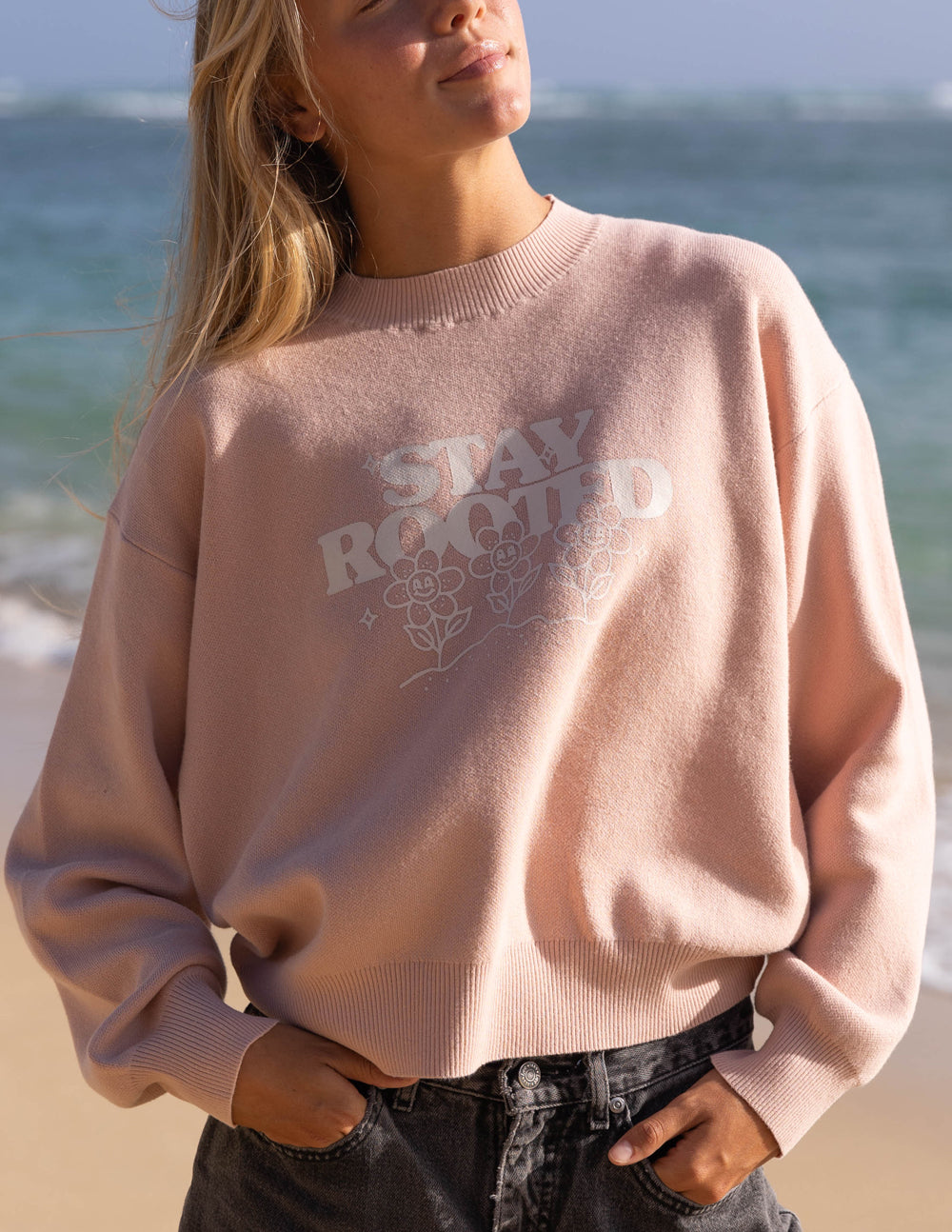 Person wearing a pink sweater with 'Stay Rooted' text on a beach